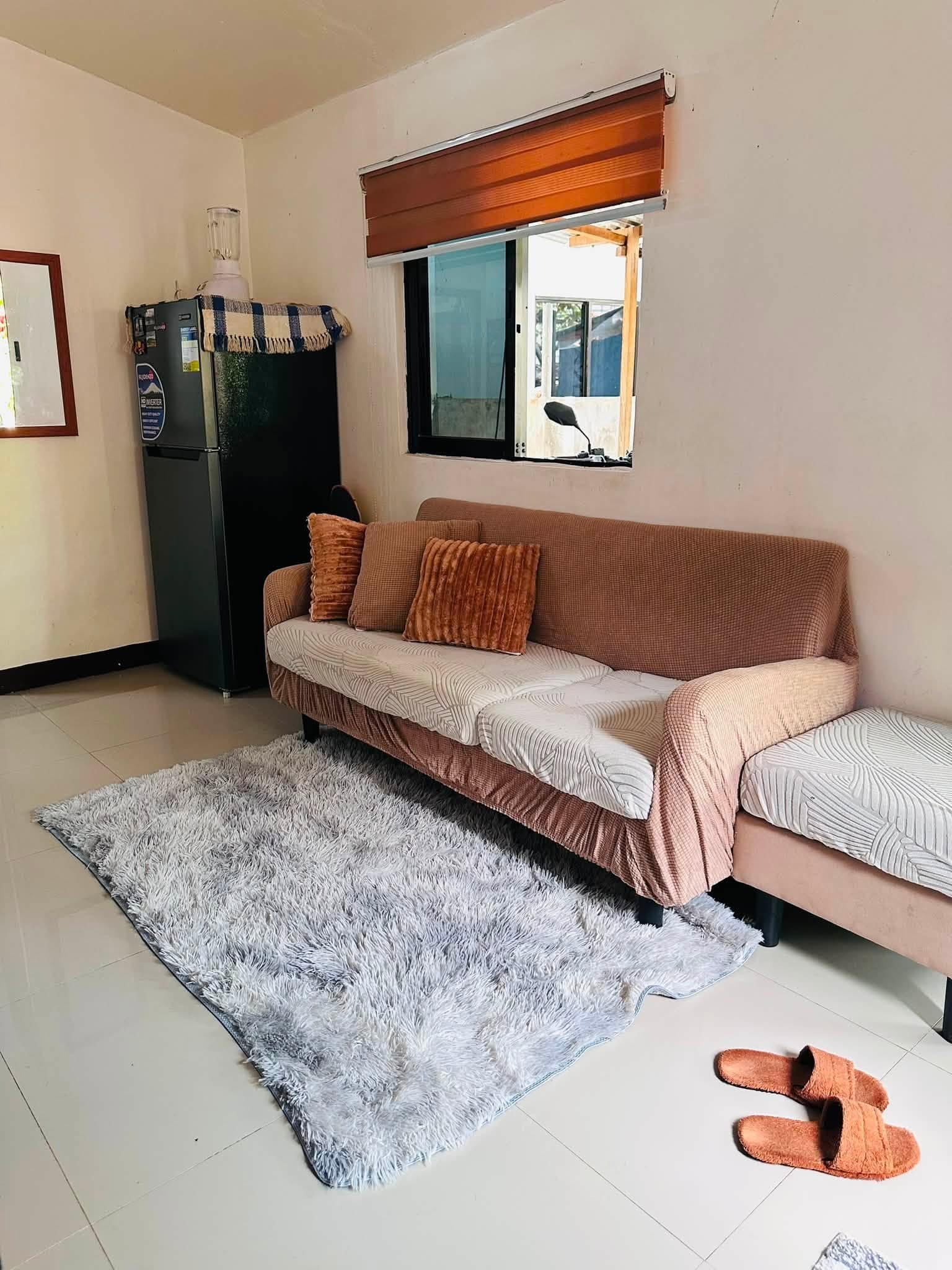 Brown sofa with rust-colored cushions on a gray shag rug, next to the refrigerator and a window with wooden blinds
