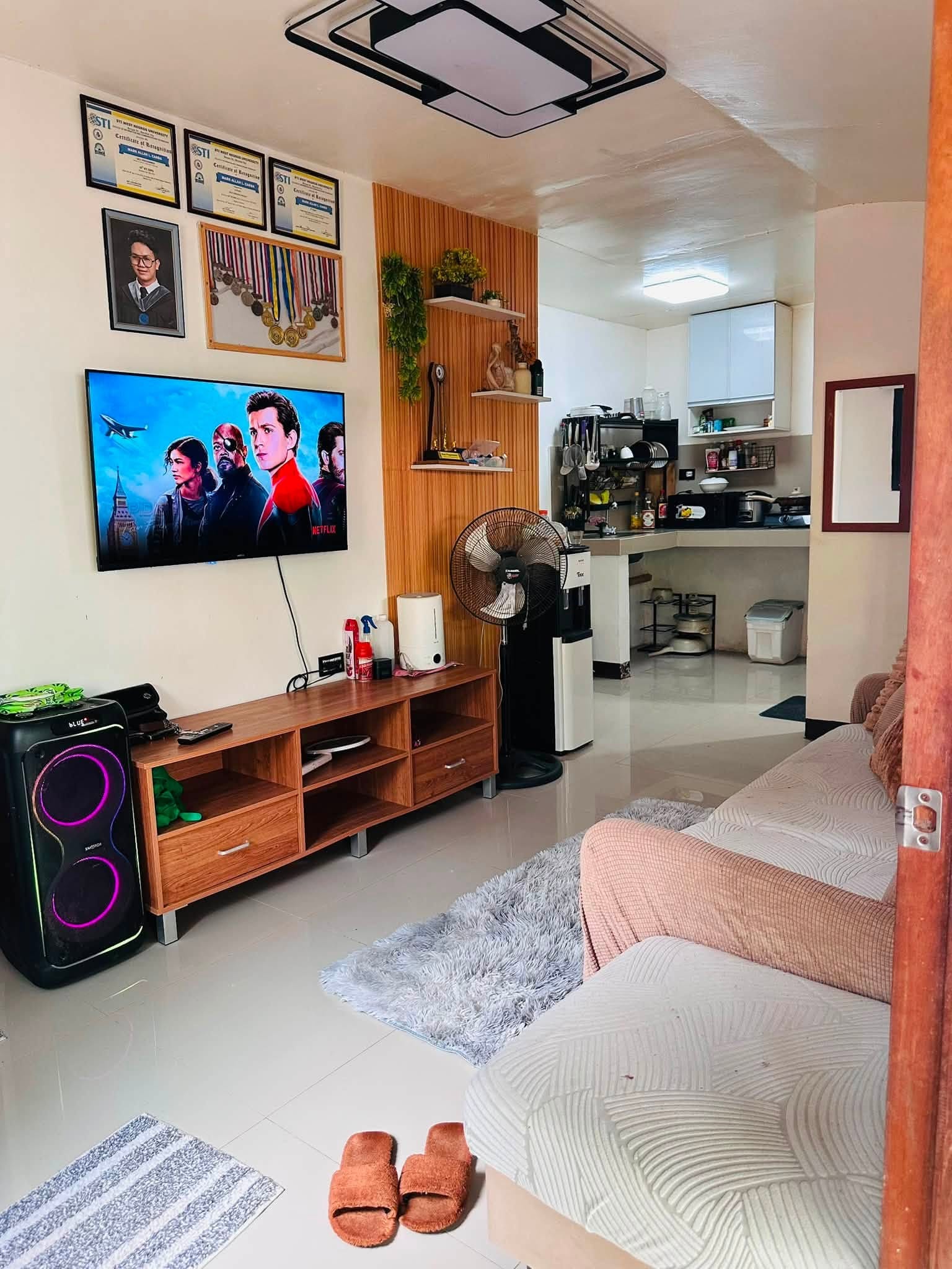 Living room with a wall-mounted smart TV showing Netflix, a wood slat accent wall, wooden TV console, karaoke speaker and a cream sofa with blush cushions, with the kitchen beyond