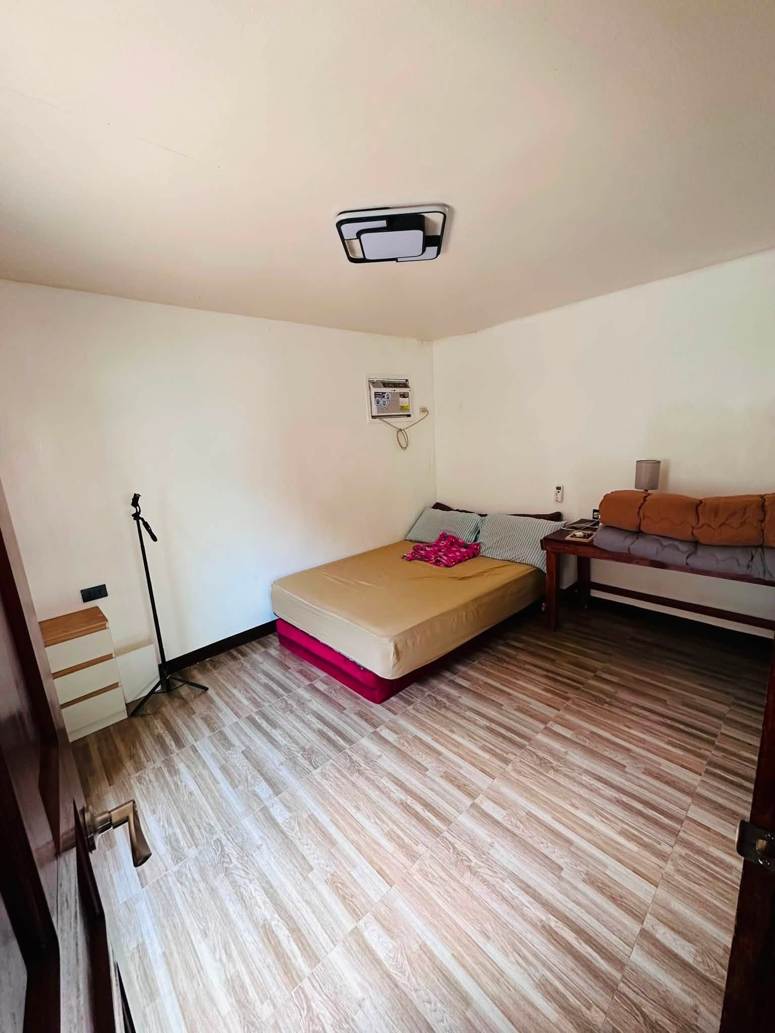 Master bedroom with a queen bed, wall air conditioner, a wooden side table with a lamp and folded blankets, and a small dresser